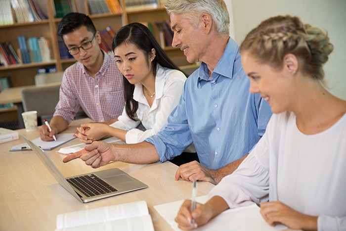 Professeur expliquant une notion à trois étudiants autour d’un ordinateur portable en bibliothèque
